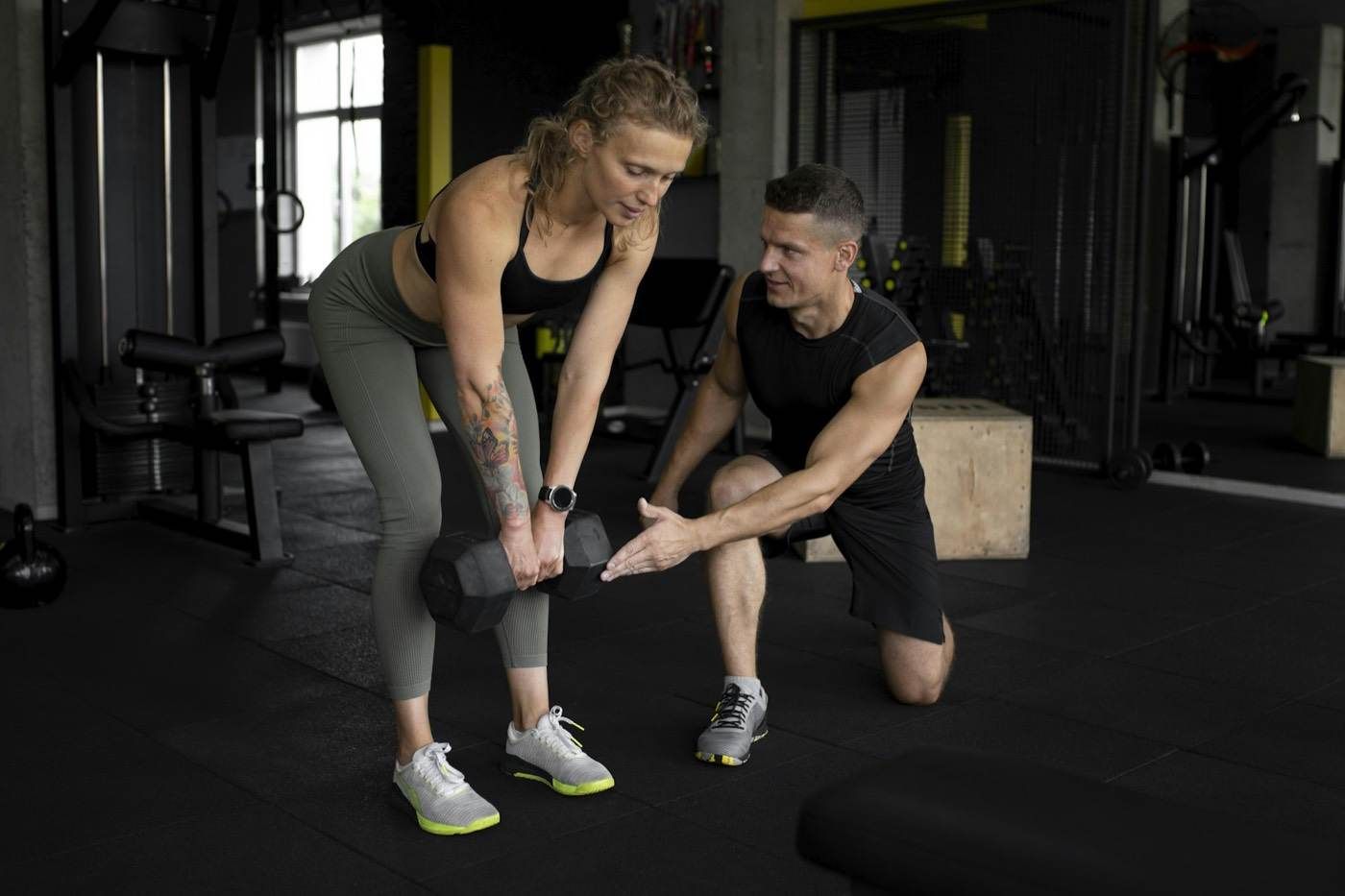 Un coach guide une cliente pendant un exercice avec haltère, dans une salle équipée de charges libres.