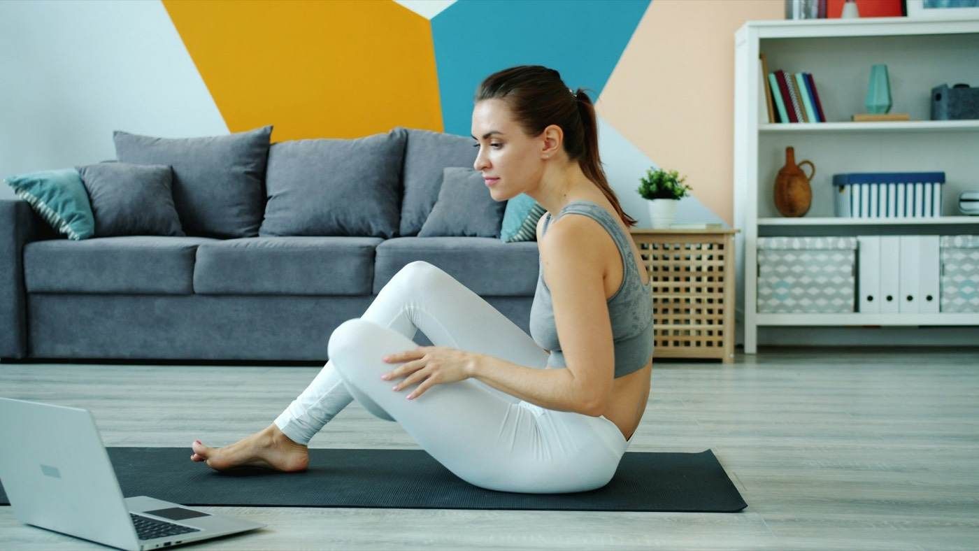 Une femme réalise un exercice de gainage assis, jambe tendue, dans un salon.