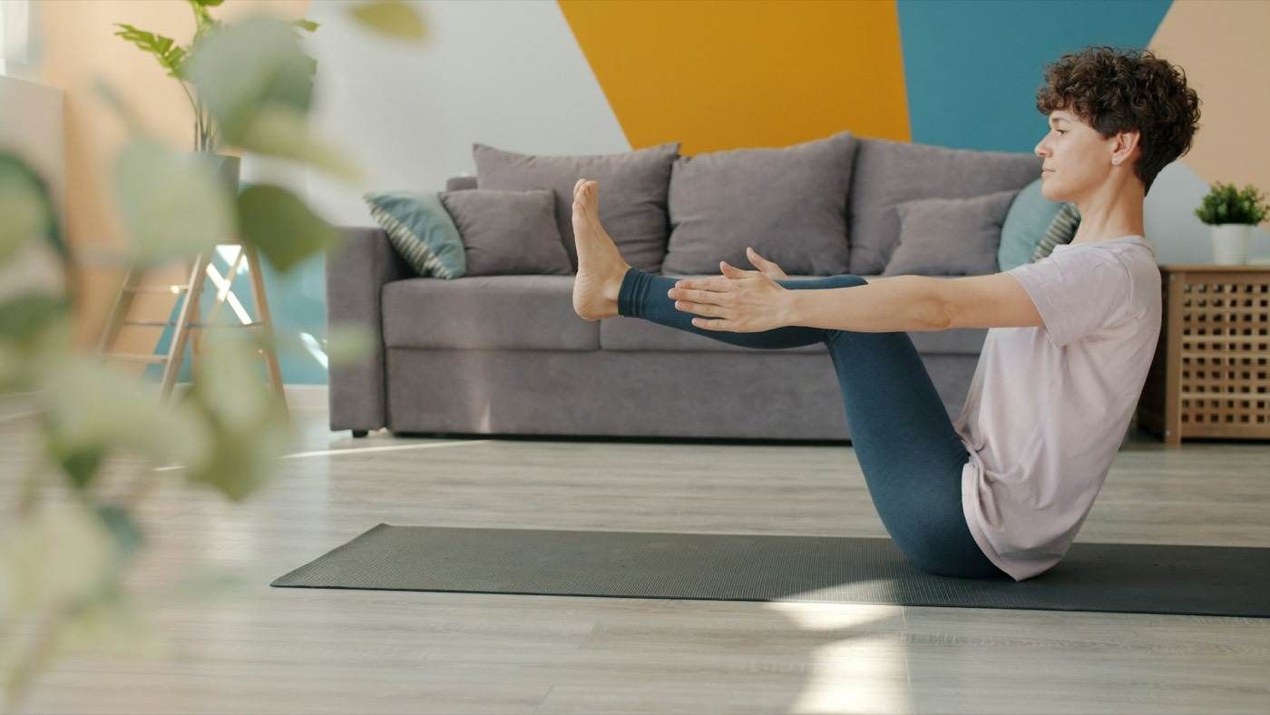 Une femme réalise un gainage abdominal assis, sur un tapis, dans un salon.