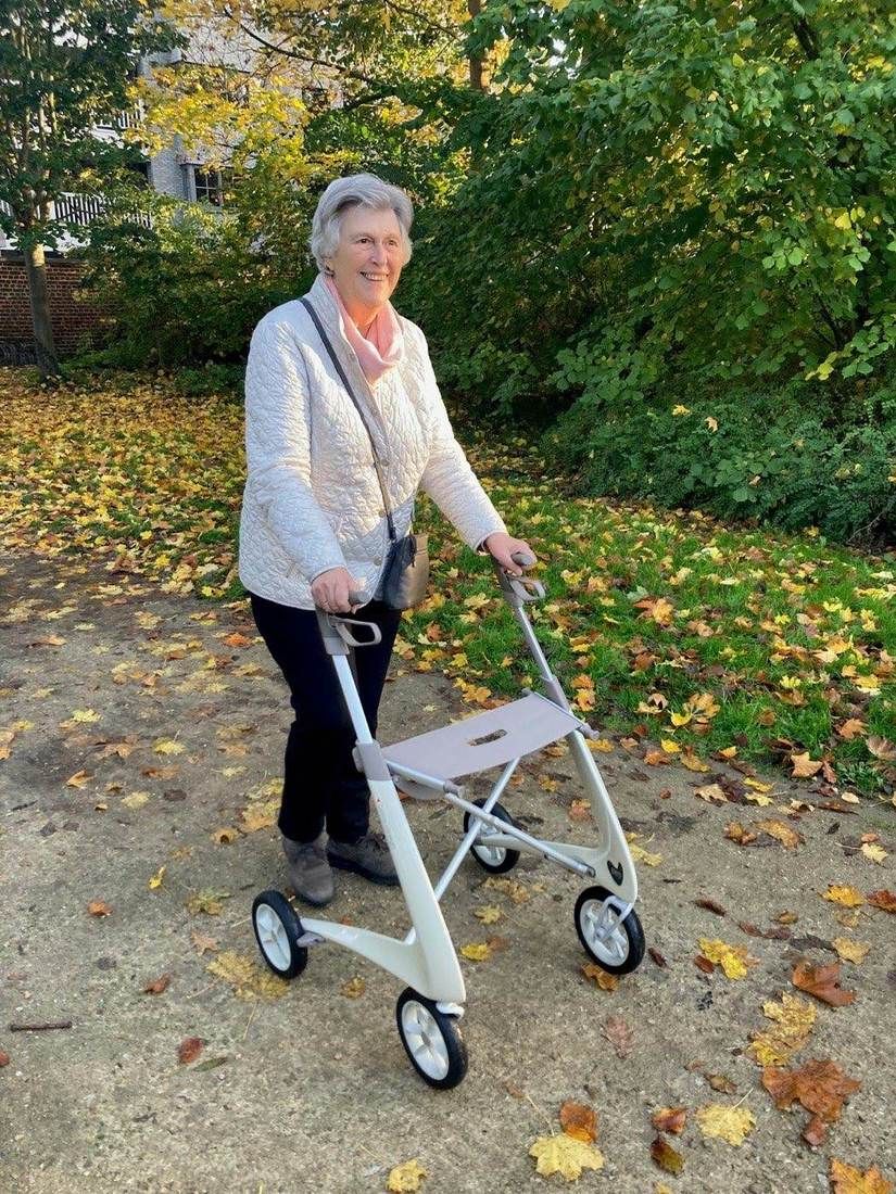 Une femme senior marche en extérieur avec un déambulateur à roulettes, parmi les feuilles d’automne.
