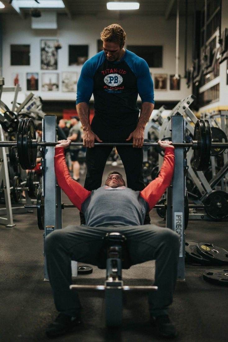 Un client réalise un développé couché avec barre, assisté par un coach, en salle de musculation.